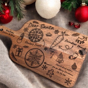 May include: Wooden Christmas serving board with engraved holiday illustrations and text. The board features a snowman, snowflake, gift, carrot, reindeer, and the words "Dear Santa," "Santa's Treat," and "Reindeer snack."