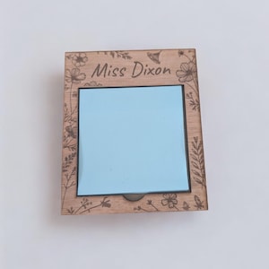Puede incluir: Un portanotas de madera con un bloc de papel azul claro. El soporte está grabado con diseños florales y el nombre "Miss Dixon". El bloc de notas es cuadrado y tiene un borde negro.