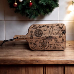 May include: A wooden cutting board with a handle, engraved with Christmas-themed illustrations and the words "Dear Santa." Includes a snowflake, snowman, carrot, reindeer, and gift box. A black cord is attached to the handle.