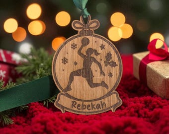 May include: Wooden Christmas ornament featuring a basketball player silhouette. The ornament is engraved with the name "Rebekah" and festive details. It hangs from a green ribbon against a blurred background with Christmas lights and a red surface.