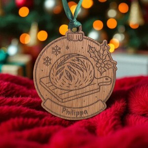 May include: Wooden Christmas ornament with a bread loaf design, snowflakes, and holly. The ornament is round with a green ribbon for hanging. The name "Philippa" is engraved at the bottom. The ornament is on a red surface.