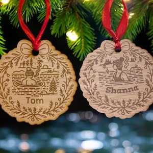May include: Two wooden Christmas decorations with red ribbons hang from a green tree. Each ornament has a scalloped edge and a laser-etched design of a person in a hot spring. One says "Tom" and the other says "Shanna".