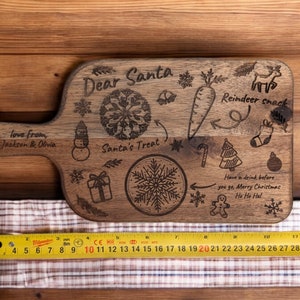 May include: A wooden cutting board with a handle, engraved with Christmas-themed illustrations and text. The text includes "Dear Santa," "Reindeer snack," and "Santa's Treat." The board also features a carrot, snowflake, and gift box design. The board measures approximately 12 inches long.