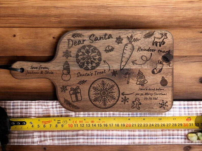 May include: A wooden cutting board with a handle, engraved with Christmas-themed illustrations and text. The text includes "Dear Santa," "Reindeer snack," and "Santa's Treat." The board also features a carrot, snowflake, and gift box design. The board measures approximately 12 inches long.
