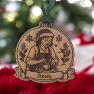 May include: Wooden Christmas bauble depicting a woman at a pottery wheel, wearing a Santa hat and apron. The bauble has a green ribbon for hanging and the name "Stacey" on a banner. The background is blurred with Christmas lights.