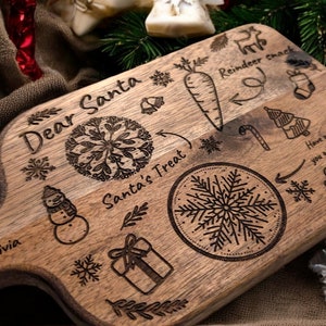 May include: A wooden serving board with engraved Christmas-themed designs. The board features text that reads "Dear Santa" and "Santa's Treat", along with illustrations of a carrot, snowflake, snowman, and gift. The board is a light brown color.