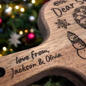 May include: Wooden serving board with a handle, engraved with the words "Dear Santa" and "love from Jackson & Olivia." The board features a snowman, snowflakes, and a decorative wreath design. A black leather strap is attached to the handle.