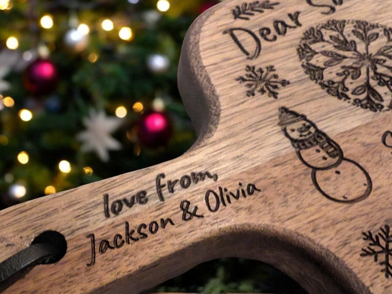 May include: Wooden serving board with a handle, engraved with the words "Dear Santa" and "love from Jackson & Olivia." The board features a snowman, snowflakes, and a decorative wreath design. A black leather strap is attached to the handle.