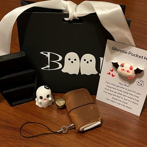 Puede incluir: Un juego de regalo temático de Halloween con una bolsa de regalo negra con "BOO" e ilustraciones de fantasmas, un pequeño ataúd, una figura de fantasma, un cuaderno de cuero marrón y una tarjeta "Ghostie Pocket Hug".