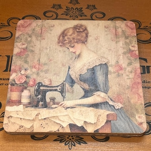 May include: A square wooden coaster featuring a vintage-style illustration of a person in a blue dress using a black sewing machine. The background displays soft pink roses and lace patterns, creating a detailed scene of sewing activity.