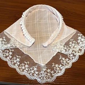 May include: A delicate, white lace collar with a soft pink backing. The collar features a rounded neckline with small, white pom-pom trim and a square bib with intricate lace edging. The fabric has a subtle plaid pattern.