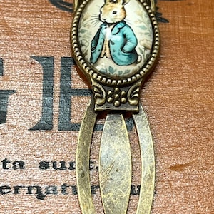 May include: Antique brass-toned bookmark featuring a detailed oval pendant with a charming illustration of a rabbit in a blue jacket. The bookmark has an elongated, openwork design, perfect for book lovers.