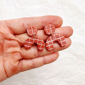 Puede incluir: Un par de pendientes de arcilla roja terracota con un patrón de cuadrícula blanca. Los pendientes tienen forma de círculo con un arco curvo debajo.