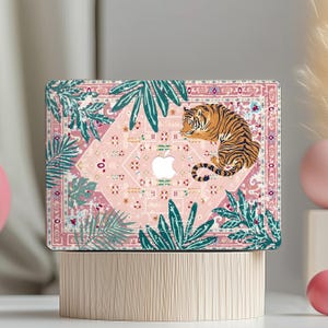 May include: A laptop skin featuring a light pink background with a decorative rug-inspired pattern, dark green tropical palm leaves, and a resting tiger. The design incorporates a central white Apple logo cutout and intricate geometric motifs.