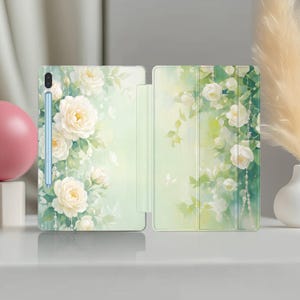 Pode incluir: Capa para tablet com um design floral de rosas brancas e borboletas sobre um fundo verde claro. A capa está aberta, revelando o ecrã do tablet e uma caneta stylus. A estética geral é suave e elegante.