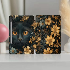 Pode incluir: Uma capa para tablet com um gato preto de olhos laranja brilhantes rodeado por flores. A capa é escura com padrões florais dourados, azuis e amarelos. A caneta do tablet é visível à esquerda.