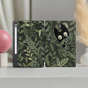 May include: A black tablet case featuring a dark green foliage pattern with various leaf shapes. A stylised black cat with large white eyes is visible peeking through the leaves on the right side. A white stylus is docked in the side slot.