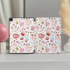 Può includere: Una custodia bianca per tablet con un motivo ripetuto di mucche rosa, fragole, cartoni di latte e stelle. La custodia è aperta, rivelando il design interno ed esterno. Il design utilizza una combinazione di colori rosa e bianco.