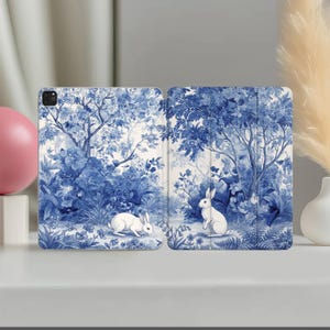 May include: A tablet case featuring a blue and white toile-style pattern. The design depicts two white rabbits in a lush forest setting with detailed trees, flowers, and foliage. The case is shown in an open, flat position, displaying the continuous illustration across both panels.