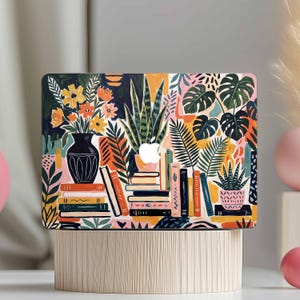 May include: A colorful laptop case featuring a vibrant illustration of books, plants, and flowers. The design includes a black vase with yellow and orange flowers, stacks of books, and various green plants. The case has an Apple logo.