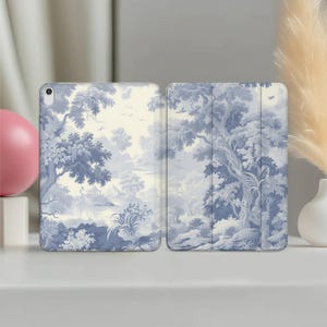 May include: A blue and white tablet case featuring a scenic landscape design. The design depicts trees, water, and birds in a classic toile style. The case is open, showcasing the interior and exterior design.