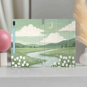 Può includere: Una custodia per tablet con un design paesaggistico. La custodia è verde chiaro e presenta un fiume che serpeggia attraverso colline verdi sotto un cielo nuvoloso con una luna crescente. Tulipani bianchi in primo piano.