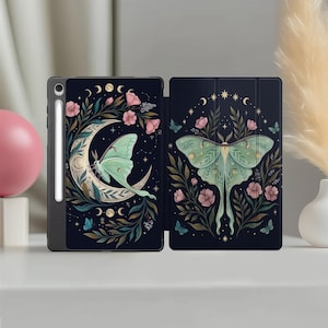 Può includere: Una custodia nera per tablet con un design a tema natura. La custodia presenta una luna crescente, una falena luna, fiori rosa e fogliame verde. La custodia aperta rivela lo schermo del tablet e uno stilo.
