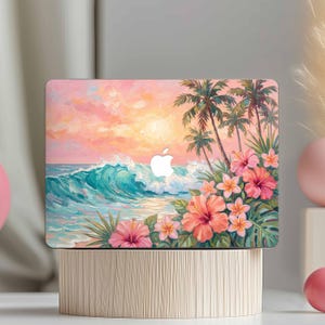 May include: Laptop case with a tropical beach design. It shows a sunset, turquoise waves, palm trees, and pink hibiscus flowers. The Apple logo is visible. The case is displayed on a wooden stand.