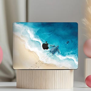 May include: Laptop cover featuring a beach scene with a sea turtle swimming in the ocean. The design includes a sandy beach, foamy waves, and a dark blue ocean. A black apple logo is visible.