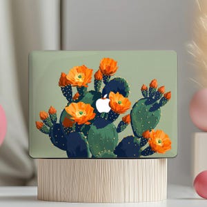 May include: A laptop case featuring a vibrant illustration of a blooming cactus. The design showcases a green cactus with orange flowers and a white Apple logo on a light green background. The case is displayed on a wooden stand.