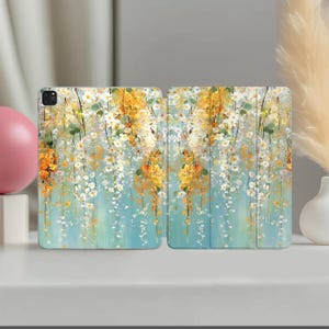 May include: A tablet case featuring a printed design of cascading white and yellow flowers against a soft teal and light blue background. The case has a tri-fold cover structure with visible vertical creases and a camera cutout on the top left.