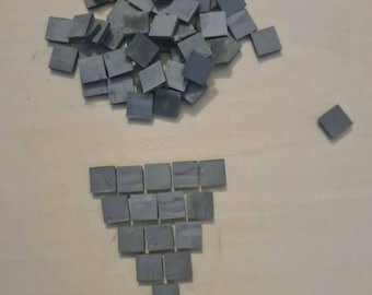 May include: A collection of small, square, gray tiles arranged in a pile and a triangle formation on a light brown surface. The tiles appear to be made of a solid material, possibly stone or ceramic. The image is well-lit, showing the details of the tiles.