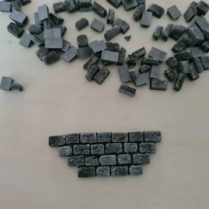 May include: A collection of small, gray, rectangular blocks, some scattered and others arranged in a brick-like pattern. The blocks have a rough, textured appearance, suggesting a stone or concrete material. The arrangement resembles a miniature wall.