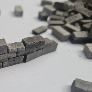 K&ouml;nnte beinhalten: Eine Miniatur-Grauziegelmauer, die aus kleinen, rechteckigen Steinen gebaut wurde. Die Mauer ist teilweise gebaut, mit einigen Ziegeln in der N&auml;he verstreut. Die Ziegel haben eine raue, strukturierte Oberfl&auml;che.