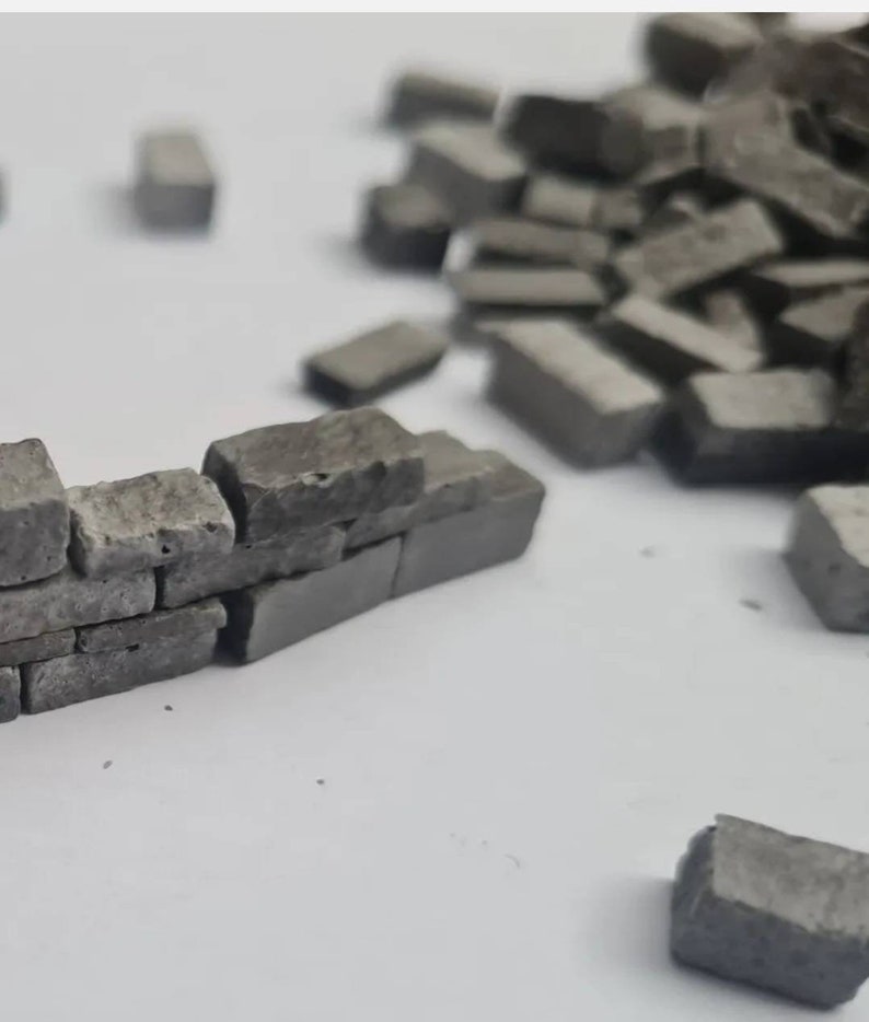 K&ouml;nnte beinhalten: Eine Miniatur-Grauziegelmauer, die aus kleinen, rechteckigen Steinen gebaut wurde. Die Mauer ist teilweise gebaut, mit einigen Ziegeln in der N&auml;he verstreut. Die Ziegel haben eine raue, strukturierte Oberfl&auml;che.