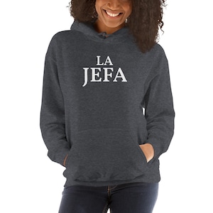 May include: Dark gray hooded sweatshirt with the white text "LA JEFA" across the chest. The sweatshirt has a front pocket and a drawstring hood. The image shows a person wearing the sweatshirt.