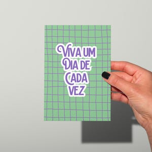 May include: A green card with a purple grid pattern and the text "Viva um dia de cada vez" in purple script. The card is held by a hand with black nail polish.