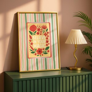 May include: A framed rectangular wall art piece featuring a vertical striped background in green, white, and pink. The center displays a yellow rectangle with red floral illustrations and the white text "BELIEVE IN YOUR MAGIC" in the middle.