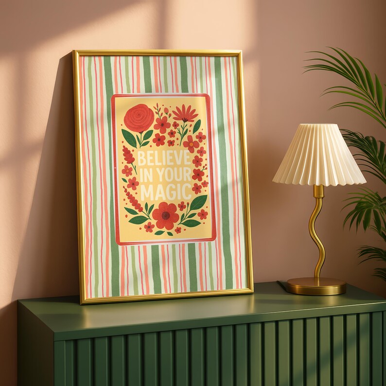 May include: A framed rectangular wall art piece featuring a vertical striped background in green, white, and pink. The center displays a yellow rectangle with red floral illustrations and the white text "BELIEVE IN YOUR MAGIC" in the middle.