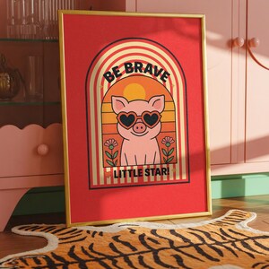 May include: A framed art print with a red background and a gold frame. The print features a pig wearing heart-shaped sunglasses, set against a retro rainbow design. Text reads "BE BRAVE LITTLE STAR!" in a playful font.