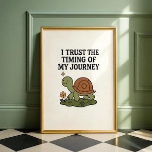May include: A framed print with a gold frame featuring a cartoon turtle and the text "I TRUST THE TIMING OF MY JOURNEY." The turtle is green with a brown shell and is standing on a patch of green grass with a flower.
