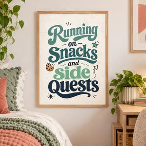 May include: A rectangular wall art print in a light wood frame featuring the text Running on Snacks and side QUESTS in dark blue and green typography. The design includes a small chocolate chip cookie icon, a star, and a curved arrow graphic.