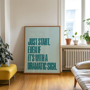 May include: A framed poster with the text "JUST START. EVEN IF IT'S WITH A DRAMATIC SIGH." The text is teal on a light blue background. The frame is a light brown color. The poster is leaning against a white wall in a room with hardwood floors.