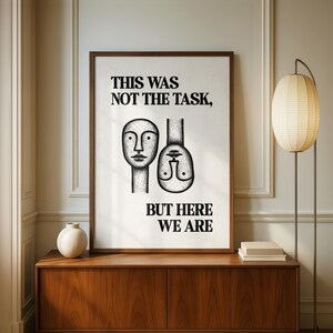 May include: A framed wall art print featuring a white background with black text that reads THIS WAS NOT THE TASK, BUT HERE WE ARE. The design includes two stylized, hand-drawn faces, one upright and one inverted, rendered in black ink lines.