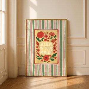 May include: A framed rectangular wall art print featuring a yellow central panel with the text BELIEVE IN YOUR MAGIC surrounded by red flowers and green leaves. The background has vertical stripes in shades of green, pink, and white.