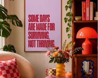 May include: A rectangular wall art print in a wooden frame featuring a light purple background with the dark pink text: SOME DAYS ARE MADE FOR SURVIVING, NOT THRIVING. The print is displayed on a wall next to a wooden bookshelf and a houseplant.