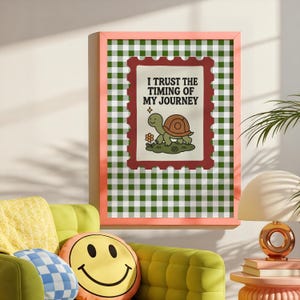 May include: A framed print with a pink border features a green and white checkered background. The print includes a postage stamp design with a turtle illustration and the text "I TRUST THE TIMING OF MY JOURNEY". The print is hanging above a green sofa.