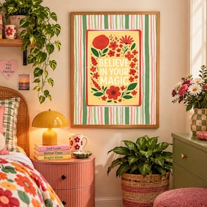 May include: A rectangular wall art print in a wooden frame featuring a yellow center panel with red flowers, green leaves, and the text "BELIEVE IN YOUR MAGIC" in white, surrounded by a border of vertical green, pink, and white stripes.