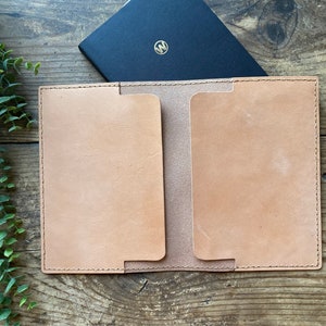 May include: A tan leather notebook cover with a stitched edge and a flap to hold a notebook. The cover is open and a black notebook is inside.