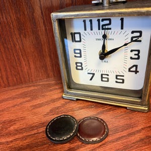 May include: Two round leather buttons, one black and one brown, with stitching around the edges. The buttons are on a wooden surface next to a vintage-style clock with a white face and black hands.